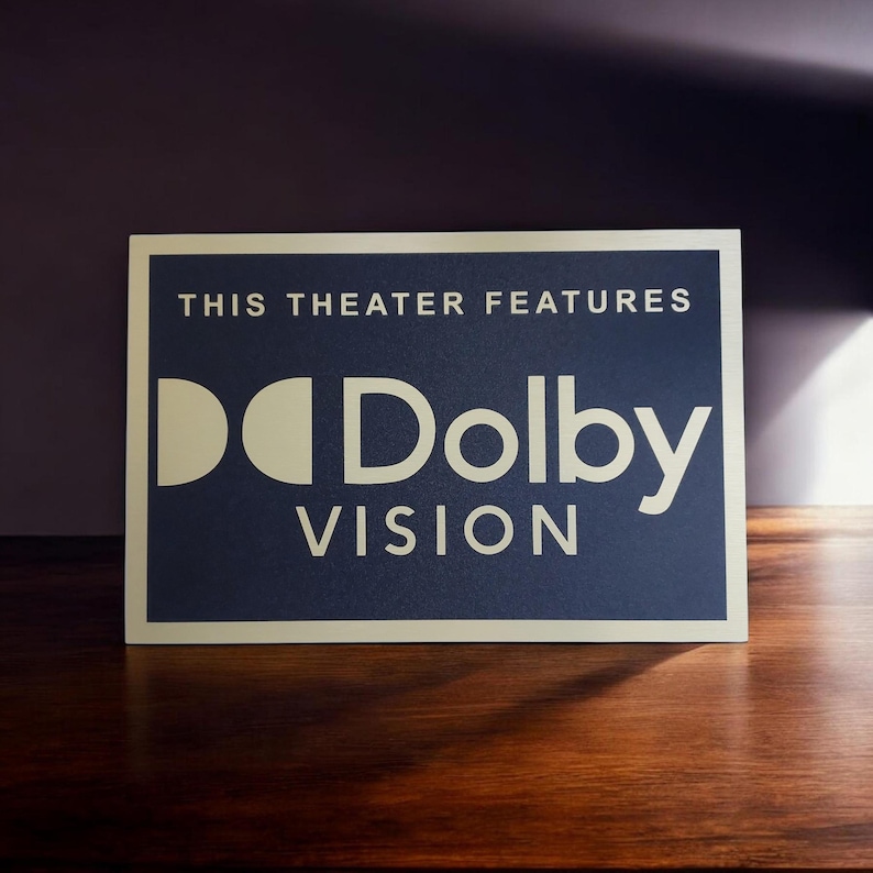 Może przedstawiać: Prostokątny znak z napisem "THIS THEATER FEATURES" nad logo "Dolby VISION". Znak ma ciemnoniebieskie tło z białym tekstem i logo. Znak znajduje się na drewnianej powierzchni.