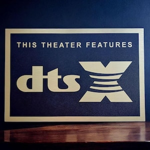 Könnte beinhalten: Ein rechteckiges Schild mit goldenem Rand und dem Text "THIS THEATER FEATURES dts X" in goldfarbenen Buchstaben. Der Hintergrund des Schildes ist dunkelblau. Das Schild liegt auf einer Holzoberfläche.