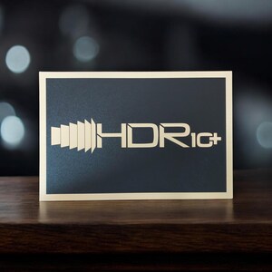 Op de afbeelding: Een rechthoekig bord met een zwarte achtergrond en een crèmekleurige rand. Het bord bevat de tekst "HDR iC+" in een crèmekleurig, gestileerd lettertype. Het bord ligt op een donker houten oppervlak.