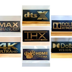 Könnte beinhalten: Eine Sammlung rechteckiger schwarzer Schilder mit goldener Metallschrift und Logos für IMAX Enhanced, DTS X, THX, 4K Ultra HD, Dolby Atmos, Dolby Vision und Home Cinema. Jedes Schild zeigt kontrastreiche Markenzeichen für Audio und Video.