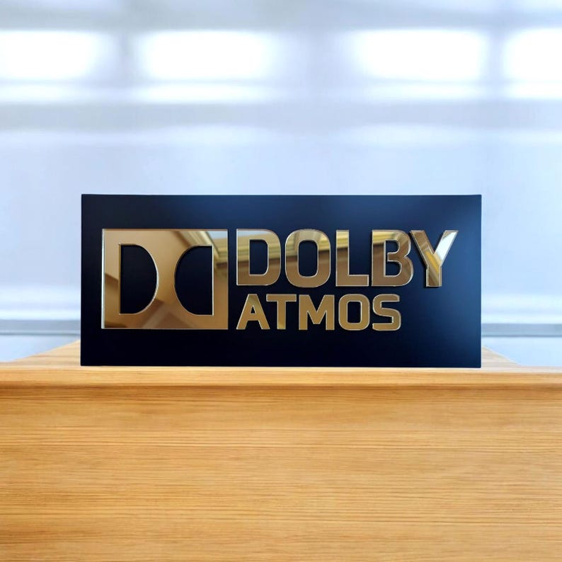 May include: A rectangular black sign featuring the gold-colored Dolby Atmos logo and text. The design includes the iconic double-D symbol on the left, with the word DOLBY stacked above the word ATMOS in a bold, metallic, reflective gold finish.