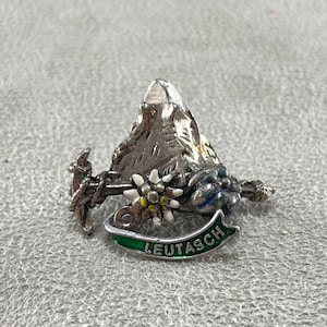 Puede incluir: Un broche de metal plateado que presenta un pico de montaña, un piolet y una flor de edelweiss con pétalos blancos y un centro amarillo. Una cinta verde curva en la parte inferior muestra el texto LEUTASCH en letras mayúsculas plateadas.
