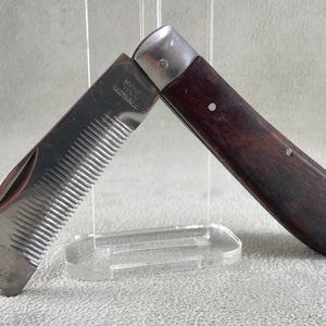 May include: A folding pocket comb with a dark brown wooden handle and a stainless steel metal comb attachment. The metal part features engraved text that reads "WARRANTED STAINLESS" and has a series of fine, parallel teeth for grooming.