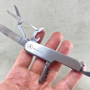 Könnte beinhalten: Ein silberfarbenes Multifunktions-Taschenmesser mit dem Mercedes-Benz Logo und Schriftzug auf dem Griff. Das Werkzeug aus poliertem Edelstahl ist ausgeklappt und zeigt eine Klinge, eine Schere, einen Flaschenöffner sowie einen Korkenzieher.