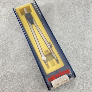 Op de afbeelding: Een zilverkleurig metalen tekenpasser met een zwarte kunststof kop met het rode rotring-logo, geplaatst in een doorzichtige kunststof etui met een gele binnenkant. De etui toont het rode rotring-logo en een wit label met 20 RS.