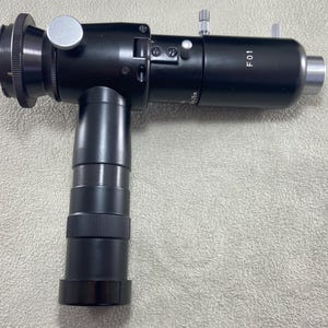 May include: A black metal colposcope featuring a T-shaped design with a vertical handle and a horizontal optical barrel. The device includes silver adjustment knobs, a silver eyepiece mount, and the white printed text F 01 on the side.