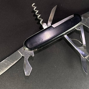 May include: A multi-tool pocket knife with a smooth black handle and multiple stainless steel implements extended, including a large blade, a corkscrew, a bottle opener, a small blade, and a pair of scissors, all arranged in a fan-like display.