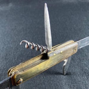 Può includere: Un coltellino svizzero multiuso con manico in ottone e diversi utensili in acciaio estratti, tra cui un cavatappi, un punteruolo appuntito e una lama principale. I componenti metallici mostrano segni di usura e invecchiamento evidenti.