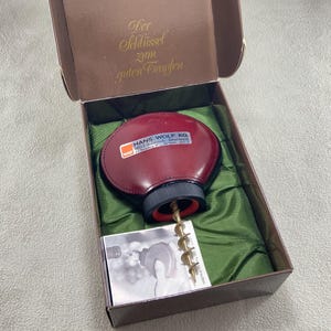 May include: A burgundy leather-covered corkscrew with a metal spiral worm, housed in a brown box with green fabric lining. The corkscrew features a label reading HANS WOLF KG 6603 Sulzbach-Altenwald. The box lid interior displays the text Der Schlüssel zum guten Tropfen.