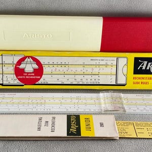 以下が含まれることがあります： 黄色と赤色のケース、計算尺、小冊子が入ったヴィンテージ計算尺セット。「ARISTO」のラベルが付いたケース。計算尺は白地に黒と黄色のマーキング。小冊子には「ARISTO JUNIOR」と記載されています。