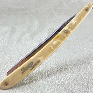 Pode incluir: Uma navalha com cabo creme e lâmina prateada. O cabo tem um acabamento perolado e é fixado com três pequenos rebites. A lâmina é gravada com as palavras "Dovo Inox".