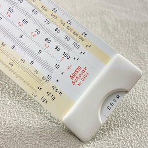 Puede incluir: Primer plano de una regla de cálculo ARISTO Scholar vintage. La regla de cálculo es blanca con marcas negras y rojas, incluyendo números, escalas y símbolos matemáticos. El cursor blanco tiene las letras DBGM impresas. Una herramienta clásica para cálculos.