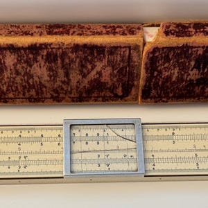 May include: A vintage off-white slide rule featuring a central sliding cursor with a clear window and black numerical markings. The instrument is accompanied by a rectangular, weathered, dark reddish-brown protective case with the text Albert Nestler.