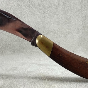 May include: A curved blade knife featuring a polished metal blade, a brass bolster, and a dark brown wooden handle secured with small brass rivets. The knife is positioned horizontally and is supported by a clear acrylic stand for display.
