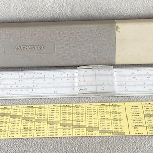 May include: A white Aristo slide rule with a clear plastic cursor, accompanied by a two-toned gray and beige protective case and a yellow Aristo conversion table card featuring various scientific units and numerical values printed in black text.