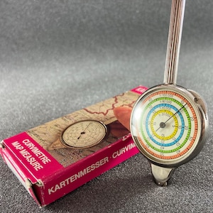 May include: A silver-toned map measure tool with a circular dial featuring concentric rings in yellow, blue, green, and orange, each with distinct scale ratios. It is positioned next to a rectangular box labeled CURVIMETRE MAP MEASURE KARTENMESSER.