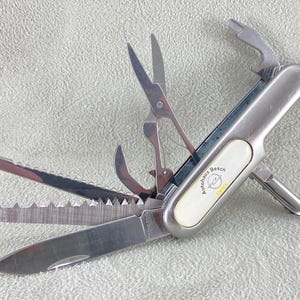 以下が含まれることがあります： ブレード、のこぎり、はさみ、ドライバーなど、複数のステンレス製ツールが展開されたシルバーのマルチツールポケットナイフ。ハンドルには、Autohaus Beschの文字とオペルのロゴが記された楕円形の白いインレイが施されています。