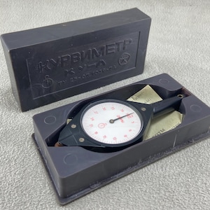 Op de afbeelding: Een zwarte mechanische kaartmeter met een witte ronde wijzerplaat met rode cijfers en een zwarte naald. Het apparaat zit in een donkergrijze kunststof behuizing met een bijpassend deksel met de in reliëf aangebrachte tekst KURVIMETR.