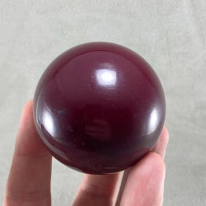 Könnte beinhalten: Eine glatte, polierte, tiefburgunderrote Kugel mit einer glänzenden, reflektierenden Oberfläche. Das runde Objekt hat eine gleichmäßige dunkelrote Farbe und ein solides, nahtloses Aussehen, das Licht auf seiner Wölbung reflektiert.