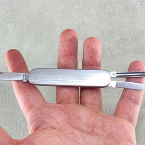 以下が含まれることがあります： 滑らかな金属製のハンドルを備えたシルバーカラーのマルチツールポケットナイフ。このナイフには、尖ったメインブレード、小さなセカンダリブレード、そしてハンドルの反対側から伸びる細い溝付きの金属製ツールが装備されています。
