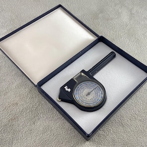 Puede incluir: Un curvímetro K&R negro con un dial circular que presenta escalas numéricas en azul y blanco, alojado en un estuche de presentación azul oscuro con bisagras y forro interior blanco. El dispositivo incluye una pequeña rueda en la base.
