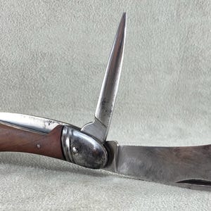 Op de afbeelding: Een inklapbaar zakmes met een houten handvat en twee metalen messen. Het ene mes is recht en puntig, terwijl het andere gebogen en breder is. Het handvat is bevestigd met kleine metalen klinknagels en heeft een metalen lus aan het uiteinde.