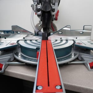 May include: A mitre saw featuring a circular metal base with a degree scale, a bright red central insert, and silver metal fences. The tool includes a dark blue motor housing, a black blade guard, and various adjustment knobs and levers.