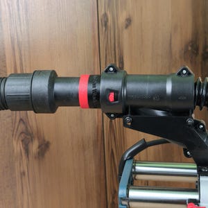 May include: A black plastic vacuum hose attachment featuring a bright red ring and a small red release button. The cylindrical component has visible screws and a textured surface, connected to a metallic structure with a red knob at the bottom.