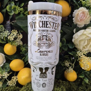 May include: A white travel tumbler with gold accents and the word "Winchester" in gold lettering. The tumbler features the text "Brothers - Hunters - Legends" and "Lawrence Kansas". The bottom of the tumbler has the text "Join the Hunt" and "Know Your Signs".