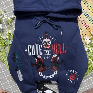Puede incluir: Una sudadera con capucha azul marino que presenta un bordado de un cordero blanco con cuernos rojos, el texto "CUTE AS HELL", dos manos enguantadas en negro sosteniendo una cadena blanca, una daga en la manga izquierda y un ojo rojo a la derecha.