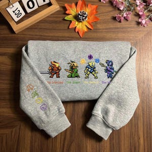 May include: A heather gray sweatshirt featuring pixelated character designs with the text "The Ironclad", "The Silent", "The Defect", and "The Watcher". The left sleeve has embroidered symbols. The sweatshirt is folded on a wooden surface.