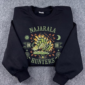 Puede incluir: Una sudadera negra de manga larga con un estampado gráfico de una criatura verde y amarilla. El texto NAJARALA está impreso sobre la criatura y HUNTERS debajo, con los números 20 y 13 a los lados, rodeados de iconos de sol y luna.