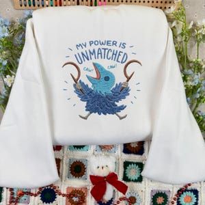 Op de afbeelding: Witte sweatshirt met de geborduurde tekst "MY POWER IS UNMATCHED" boven een blauw vogelontwerp met bruine sikkels. De vogel heeft een blauw lichaam en een blauwe snavel. Het woord "CAW!" is aan weerszijden van de vogel geborduurd.