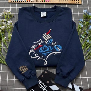 Puede incluir: Una sudadera azul marino de cuello redondo con un bordado central de una mano esquelética sosteniendo un gancho rojo sobre una rosa azul y detalles blancos. La manga derecha tiene un logotipo dorado bordado y una etiqueta blanca en el cuello.