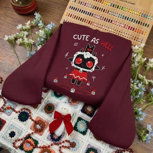 Può includere: Una felpa bordeaux con la frase ricamata "CUTE AS HELL" e un disegno di agnello dei cartoni animati. L'agnello ha gli occhi rossi e indossa un abito rosso. La felpa è esposta su una colorata coperta all'uncinetto.