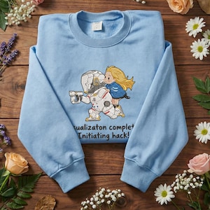 Puede incluir: Sudadera azul claro con un dibujo animado de un astronauta futurista y una niña. El astronauta sostiene un arma. El texto "ualizaton complet Initiating hack!" está impreso debajo. La sudadera tiene cuello redondo.