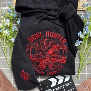 Op de afbeelding: Een zwarte hoodie met rode borduursels op de borst en mouw. Het ontwerp op de borst toont twee gekruiste pistolen, een schedel en de tekst "DEVIL HUNTER", "LET'S ROCK", "EST. 2001", "SSS RANK" en "MISSION COMPLETE".