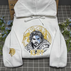 Könnte beinhalten: Ein weißer Kapuzenpullover mit einer großen Stickerei auf der Brust, die eine bärtige Person mit blauen Gesichtszügen zeigt, umgeben von einem goldenen kreisförmigen Pferdemotiv und dem Schriftzug KLIFF. Ein kleines goldenes Pferdemotiv ist am Ärmel.