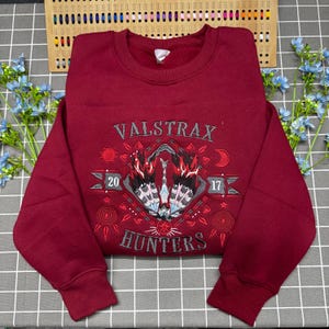 May include: A maroon crewneck sweatshirt featuring an embroidered graphic with the text VALSTRAX HUNTERS and the numbers 20 and 17. The design includes a central stylised creature motif in black, red, and white, surrounded by sun and moon symbols.