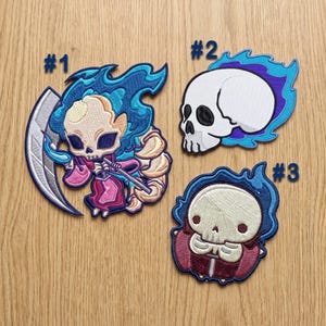 May include: Three embroidered patches labelled #1, #2, and #3. Patch #1 shows a skeleton figure with a scythe and blue flames. Patch #2 is a white skull surrounded by blue flames. Patch #3 is a small skeleton figure wearing a red cloak with blue flames.