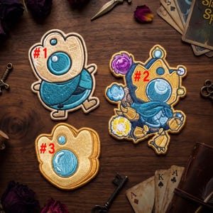 May include: Three embroidered patches on a wooden surface. Patch #1 is a tan and teal creature. Patch #2 is a complex tan, teal, and purple character with a crown. Patch #3 is a tan, shell-shaped design with two blue circular accents.