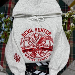 Può includere: Una felpa con cappuccio grigio chiaro caratterizzata da una stampa grafica rossa sul petto e sulla manica. Il design centrale mostra due pistole incrociate, un teschio e il testo "DEVIL HUNTER", "LET'S ROCK", "EST. 2001", "SSS RANK" e "MISSION COMPLETE".