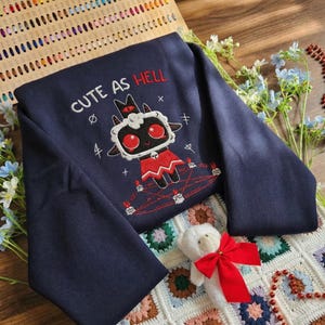 May include: Navy blue sweatshirt with the embroidered phrase "CUTE AS HELL" and a cartoon lamb design. The lamb has red eyes and a red outfit. The sweatshirt is folded on a colorful, patterned blanket.