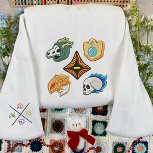 Può includere: Felpa bianca con toppe ricamate a tema fantasy. Le toppe includono un teschio, uno scudo e altre forme ornate in oro, verde e blu. La manica sinistra presenta un piccolo disegno ricamato. Un piccolo orsacchiotto con un fiocco rosso è in primo piano.