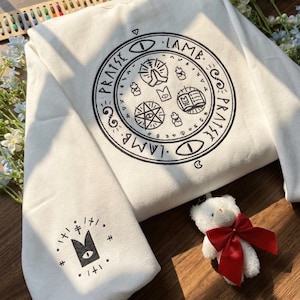 Może przedstawiać: Kremowa bluza z dużym czarnym okrągłym wzorem z przodu, z napisem "PRAISE LAMB" i różnymi symbolami. Rękaw ma mniejszą grafikę. Biały pluszowy miś z czerwoną kokardą jest na pierwszym planie.
