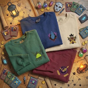 May include: Four crewneck sweatshirts in blue, green, beige, and burgundy, each with an embroidered design. The sweatshirts are arranged on a wooden surface with fantasy-themed props like cards, potions, and maps. The designs include a hand, a crown, a clock, and a plant.