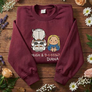 May include: A maroon sweatshirt featuring a cartoon illustration of two figures, one in a space suit and the other with blonde hair, with the names "HUGH & D-I-03367 DIANA" printed below. The sweatshirt is made of a soft material.