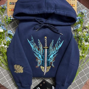 Op de afbeelding: Een marineblauwe hoodie met een grote, centrale geborduurde afbeelding van een gestileerde wezenkop met een verticaal zwaard erdoorheen in tinten lichtblauw, bruin en goud. De rechtermouw is voorzien van een goud geborduurd logo.