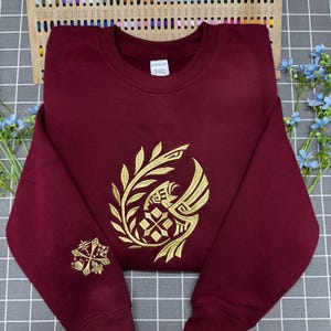 Puede incluir: Una sudadera de cuello redondo color granate con bordados dorados en el pecho y la manga izquierda. El diseño del pecho muestra un ave estilizada dentro de una corona de laurel, mientras que la manga presenta un emblema geométrico.
