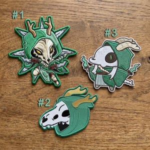 May include: Three embroidered patches featuring hooded figures with skull-like masks. Patch #1 shows a figure with antlers and blades. Patch #2 displays a profile of a skull-masked figure. Patch #3 depicts a small figure with horns and green flames.